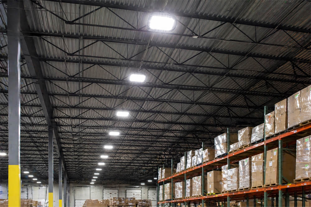 High-bay LED lighting fixtures illuminating a large warehouse ceiling with metal trusses and rows of stacked palletized boxes on industrial shelving