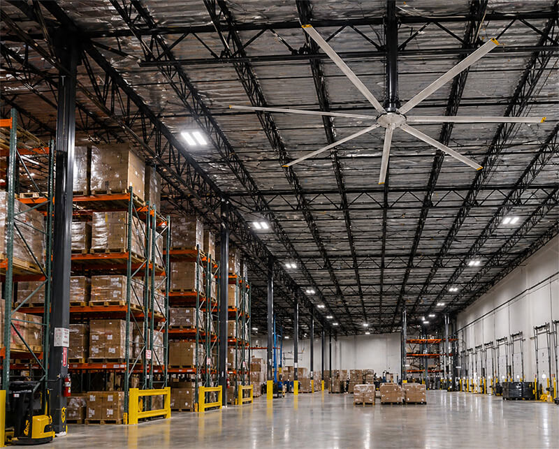 Large industrial warehouse interior with high ceilings, steel trusses, and an installed HVLS ceiling fan providing airflow across open storage and pallet areas.