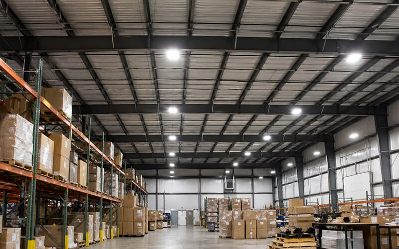 Bright LED lighting illuminating a large industrial warehouse with high ceilings, metal shelving, and organized rows of palletized inventory